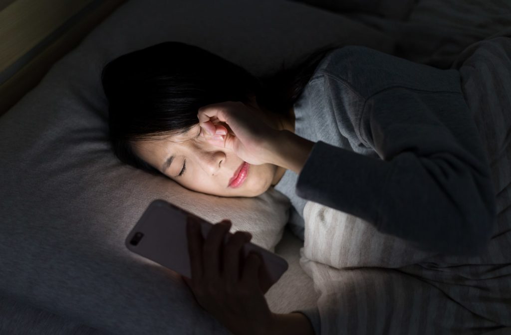 A woman rubbing her eyes while looking at her phone in bed