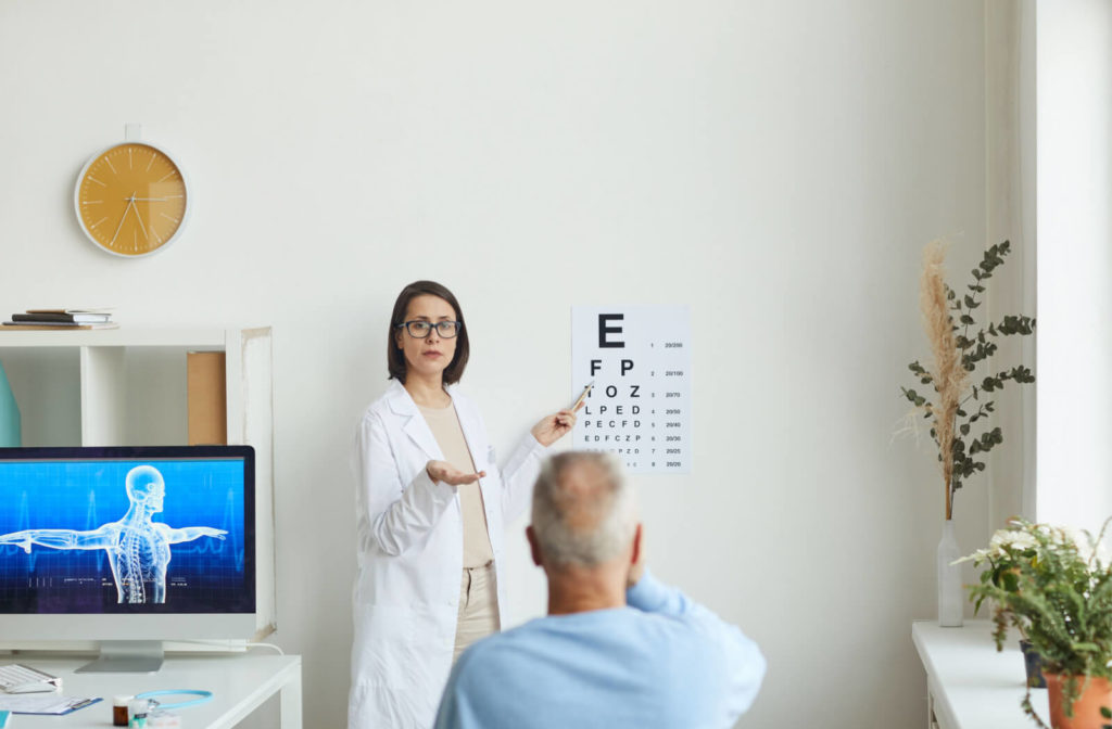 A male senior getting his regular eye exam, sitting afar from the optician and covering his right eye while the optician is ponting a letter from a Snellen chart.

