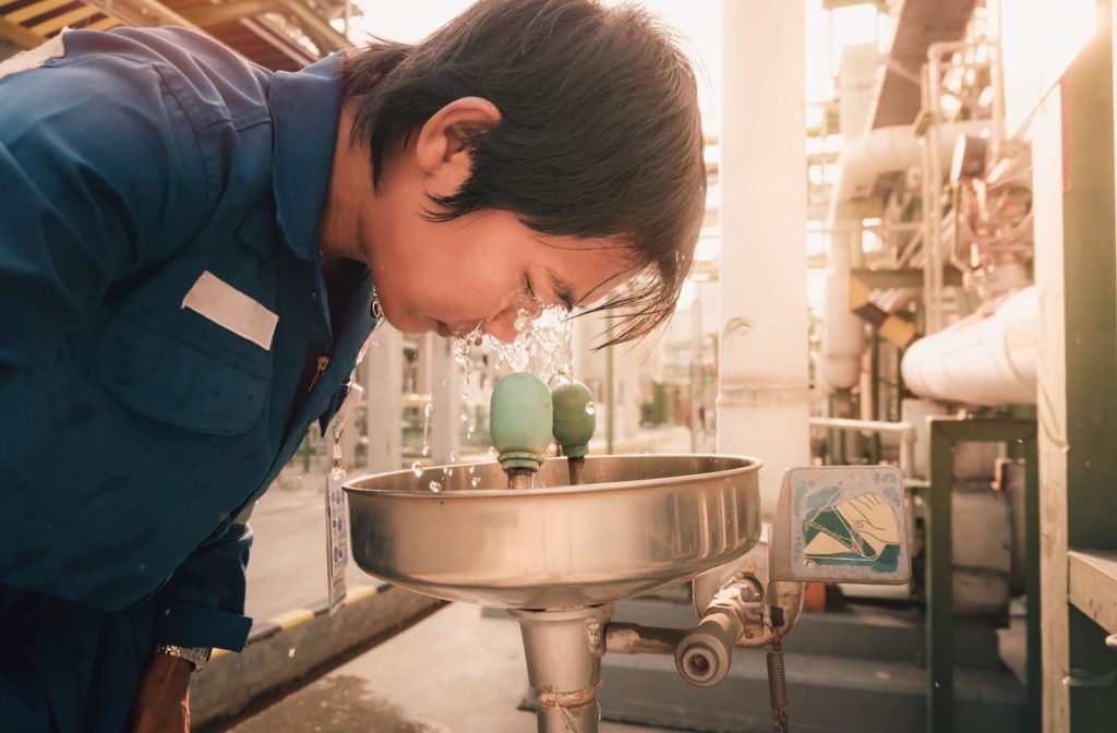 A person rinses their eyes at an emergency eye wash station. The person is wearing coveralls, and industrial equipment is visible in the background, showing that they're at work.