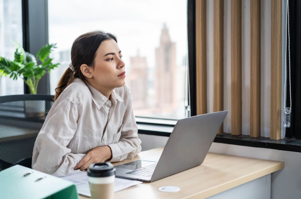 A woman sitting at her desk in front of her laptop. She is taking a break by looking away from the screen.