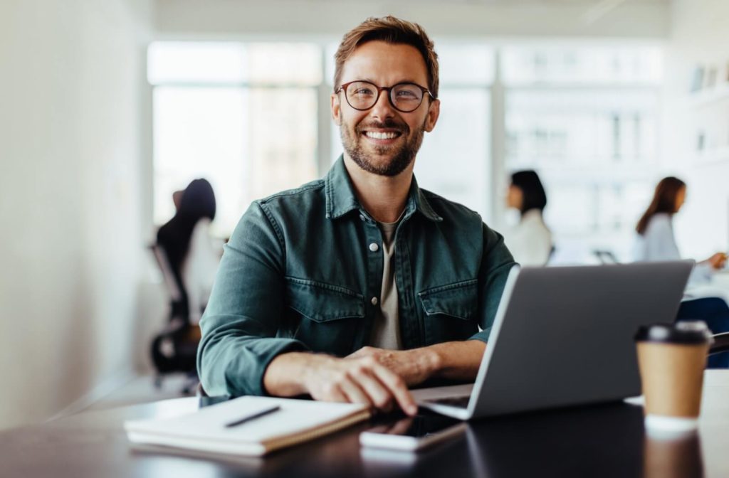 Smiling man wearing eyeglasses, sitting at a desk with a laptop, notebook, and coffee cup in the office