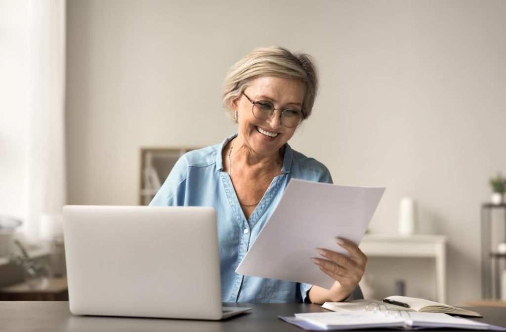 Someone wearing glasses switches between working on their laptop and paperwork.