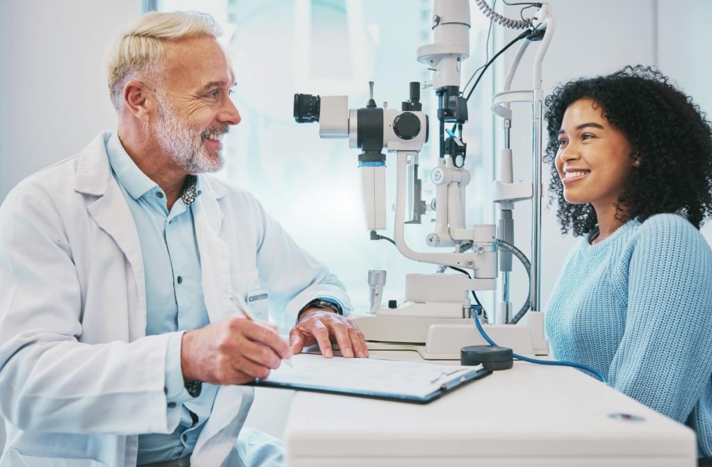 An eye care professional sits with a patient during an eye exam, smiling while taking notes beside a slit lamp machine.