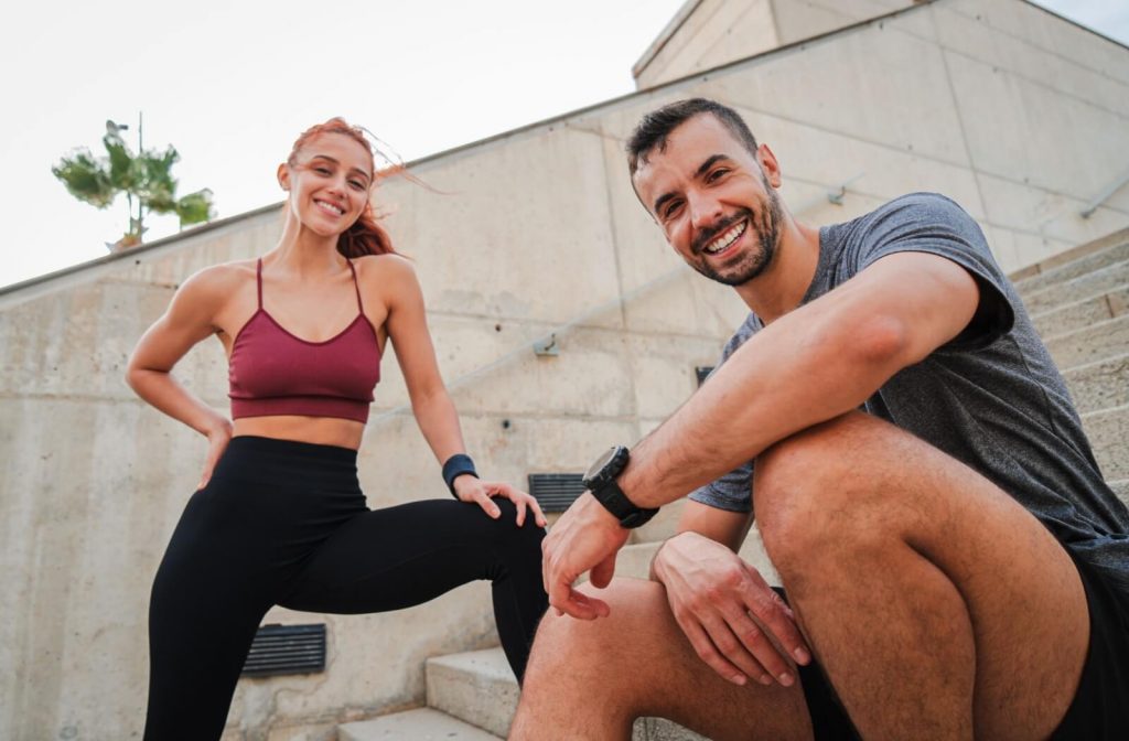 Two people in athletic wear smiling and taking a break on outdoor concrete steps after exercising.