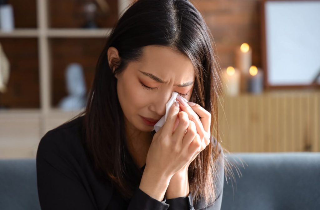 Person sitting indoors wiping their eyes with a tissue, appearing to experience stinging or discomfort while crying.