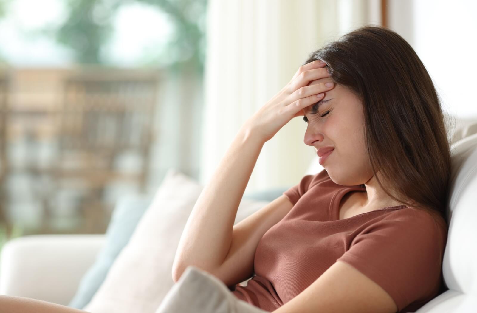 A person sitting on a couch with their eyes closed, resting one hand on their forehead as if experiencing a headache or migraine.